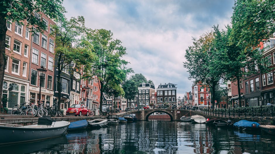 Kanał w miejskiej scenerii, który jest charakterystyczny dla Amsterdamu. Po obu stronach kanału znajdują się zabytkowe, wąskie domy z cegły, w typowym dla Holandii stylu. Na pierwszym planie, przy brzegu kanału, cumują liczne łodzie i barki. Nad kanałem przerzucony jest kamienny most z balustradą, po którym przechodzą ludzie i rowery, typowy element miejskiego krajobrazu w Holandii. Drzewa o liściach w odcieniach zieleni sugerują, że jest to zdjęcie wykonane w cieplejszej porze roku. Niebo jest częściowo pokryte chmurami, co nadaje zdjęciu atmosferę wczesnego popołudnia lub późnego poranka.